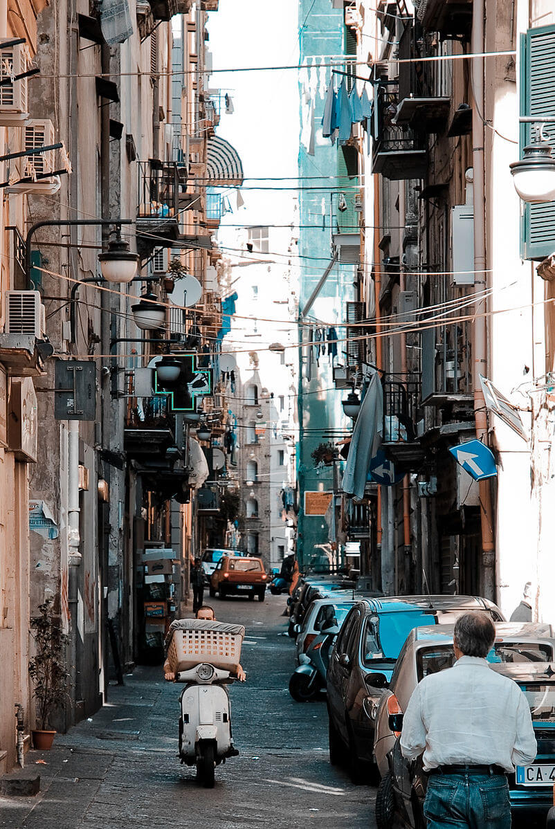 A narrow but busy street in Naples, overhung by high apartment blocks.